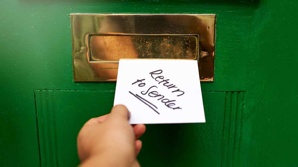 Eine Hand schiebt ein Brief-Kuvert mit der Aufschrift "Return to Sender" in einen Briefkastenschlitz in einer grünen Türe.