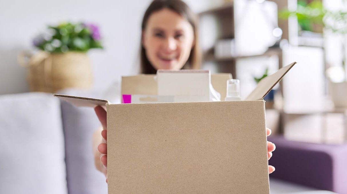 Eine lachende junge Frau hält ein Paket in die Kamera, das verschiedene Packungen mit Waren enthält.