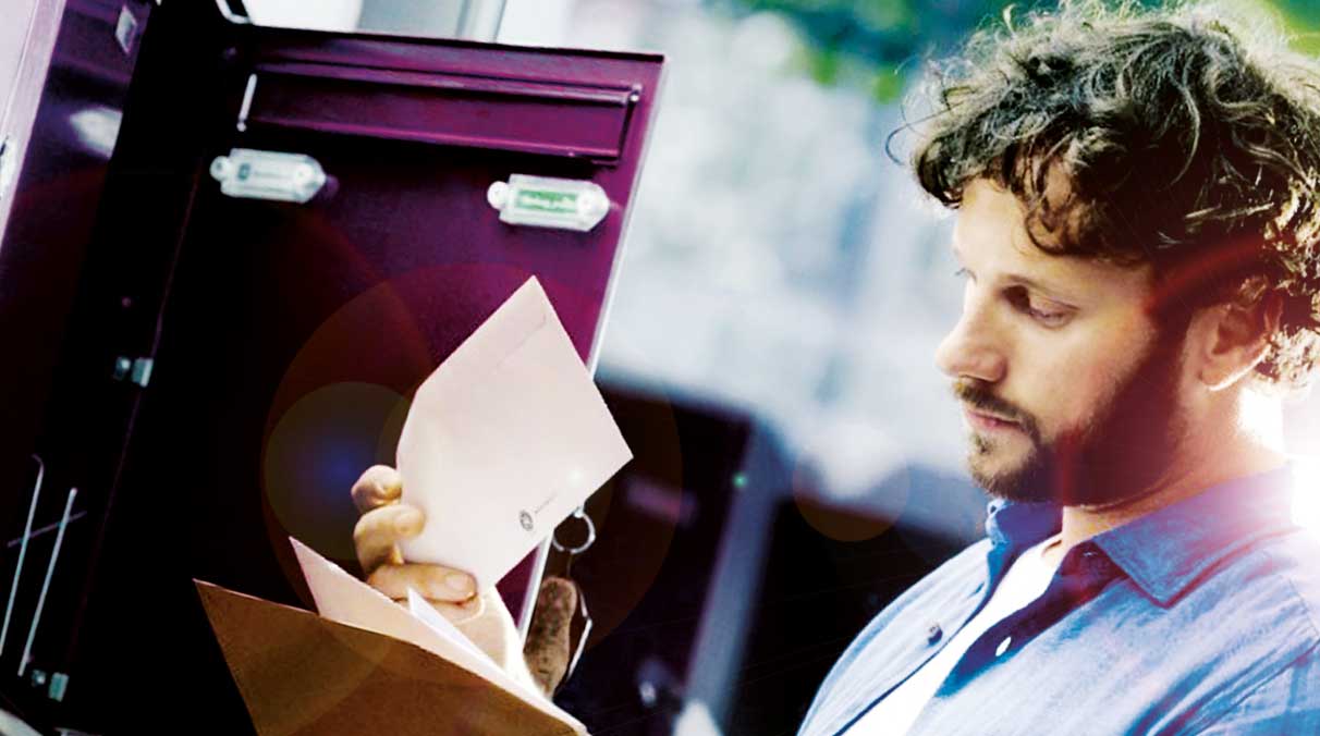 Junger Mann steht vor geöffnetem Briefkasten und hält offenes Briefkuvert in der Hand